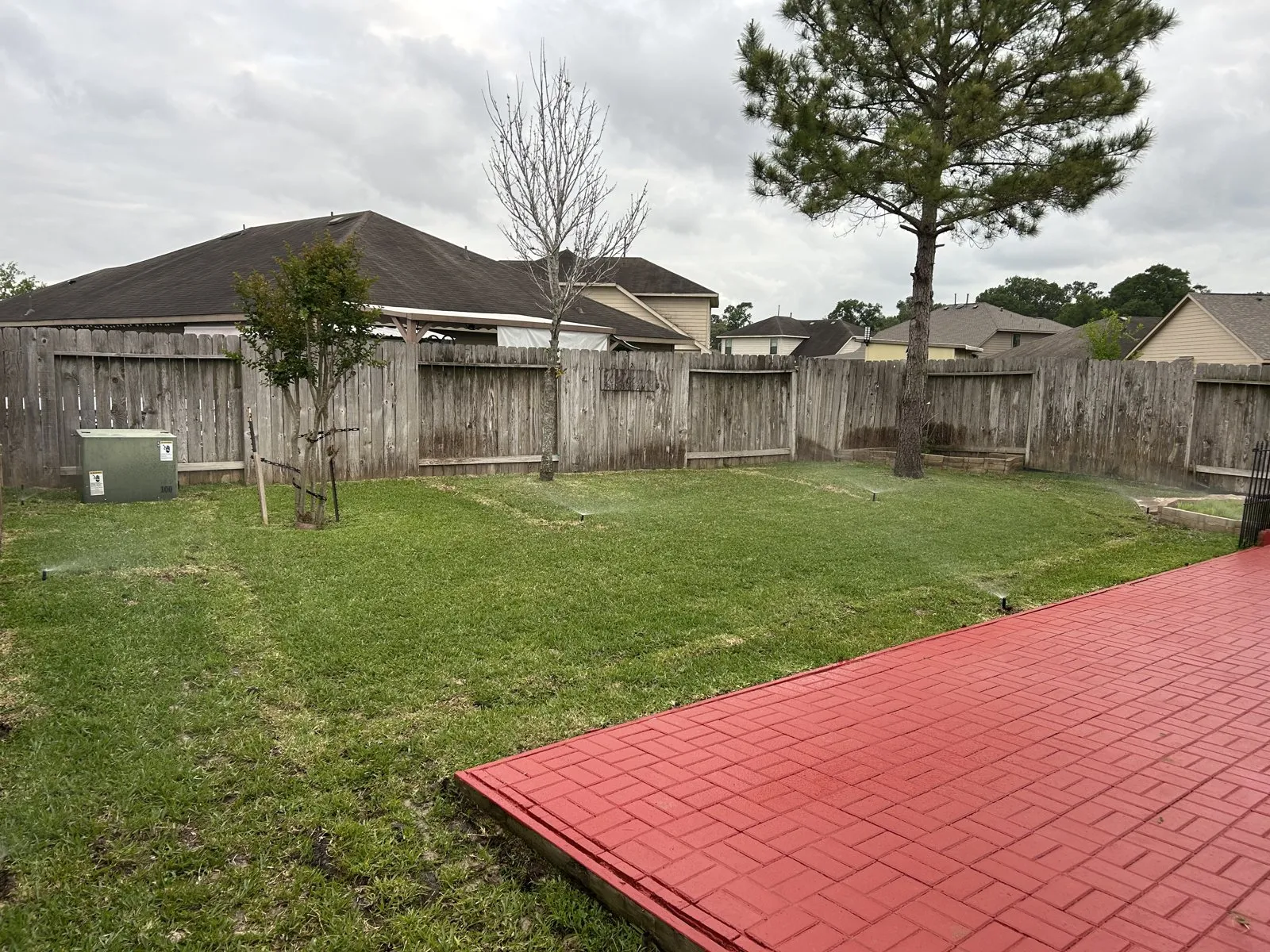 Sprinklers watering a fenced backyard beside a red patio